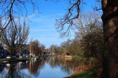 Köprüden bir kanal manzarası, bir tarafında park, diğer tarafında arabalar olan bir yol. Parlak mavi gökyüzü ve güneş doğanın Nisan ayındaki güzelliğini vurguluyor..