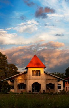 Üzerinde beyaz bir haç bulunan, parlak bir alacakaranlık gökyüzüne karşı duran kırmızı kiremitli bir kule içeren basit beton bir kilise yapısı..