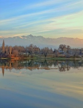Orman, dağlar ve mavi gökyüzü sakin bir göle yansıyor. Fotoğraf, doğanın atmosferini, ahengi ve resimsel manzarayı aktarır..