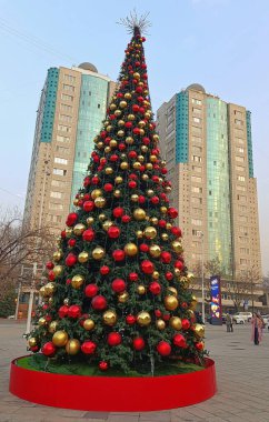 Kırmızı ve altın süslemeli süslü bir Noel ağacı şehir meydanında, uzun ve modern bir cam binanın önünde duruyor. Kentsel kış atmosferi, halka açık tatil süslemeleri. 