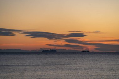 Güneş doğmadan önce seyir eden siluetli gemiler, katmanlı bulutlar ve endüstriyel gemileri çevreleyen uzak kıyı şeridi, sakin suyu yansıtan yumuşak turuncu şafak öncesi ışık, dengesiz denizcilik