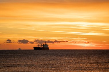 Gündoğumu gökyüzü tarafından çerçevelenmiş büyük bir taşıyıcı, sakin suya yansıyan ateşli bulut bantları, güçlü bir kontrast yaratan uzak siluet, erken endüstriyel rutin ruh hali, cömert gökyüzü alanı