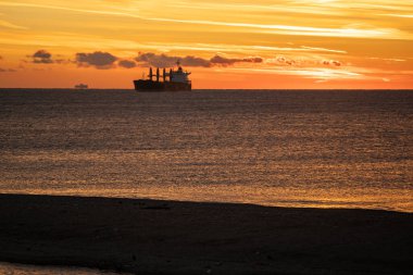 Sahil yük gemisi şafak vakti sakin körfezden geçiyor, gökyüzü ve suyun altın katmanları, alçak kıyı şeridi, ufuk çizgisinden geçen kargo silueti, yolculuk için ideal deniz sabahı