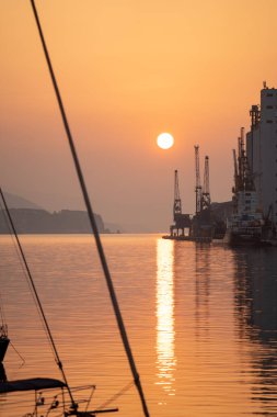 Sakin bir kanalın üzerine düşen güneşe karşı çerçevelenmiş yelkenli donanımı, liman endüstriyel şekiller parlarken ipler ve direkler siluet halinde