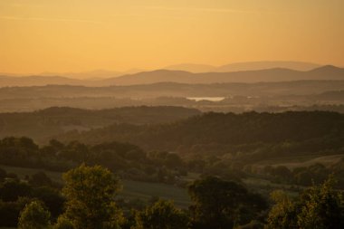 Geniş altın vadi, katmanlı tepeler ve uzak siluetler, sıcak ışık boşluklarında birikiyor.