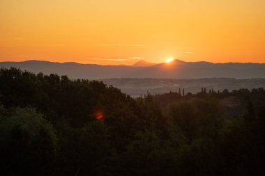 Akşam güneşi katmanlı tepelerin ardında inerken, kehribar gökyüzü uzak siluetleri taçlandırırken ön plandaki ağaçlar kalır.