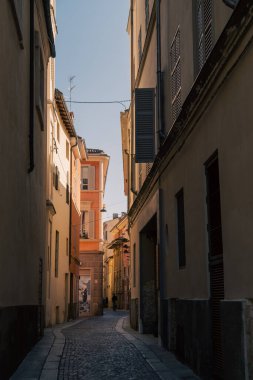 Çerçevelenmiş uzun sıva binalar, kepenkli pencereler, sıcak öğleden sonra ışığı, kapı girişinin yakınındaki yalnız figür, sessiz bir ev atmosferi.,
