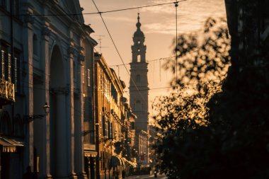 Bulvar ve kulede sıcak gün batımı aydınlatması, klasik cepheler parlıyor, tramvay hatları üstümüzde, uzun gölgeler ve uzak çan kulesi sunucu sahnesi, yoldan geçenler ve sokak detayları