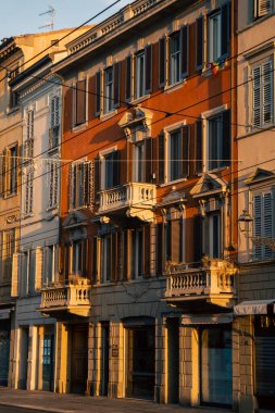 Parma cephesinde sıcak öğleden sonra ışığı, süslü balkonlar ve ahşap kepenkler uzun desenli gölgeler, zarif kornişler, soluk sıva ve dar sokak perspektifi
