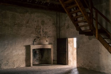 Eski şöminesi olan boş bir oda, yumuşak kapı ışığıyla aydınlatılmış bekçi odası, gölgeye yükselen ahşap merdiven, alçı ve is lekeli şömine.