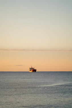 Güneş doğarken yalnız kargo gemisi, mesaj için yer. Sakin deniz ve pastel gökyüzü ufka kadar uzanıyor, minimalist kompozisyonun merkezindeki uzak siluet yalnızlığı ve yavaş denizciliği çağrıştırıyor.