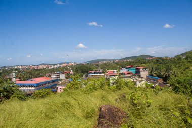 Açık mavi gökyüzünün altında renkli binalar, palmiye ağaçları ve cami minareleri olan tropikal bir Hint kasabasının panoramik manzarası.