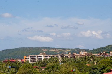 Cityscape of Goa, India featuring residential buildings, palm trees, and green hills under a clear blue sky.