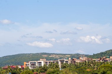Goa, Hindistan şehir manzarasında açık mavi gökyüzü altında konut binaları, palmiye ağaçları ve yeşil tepeler yer almaktadır..