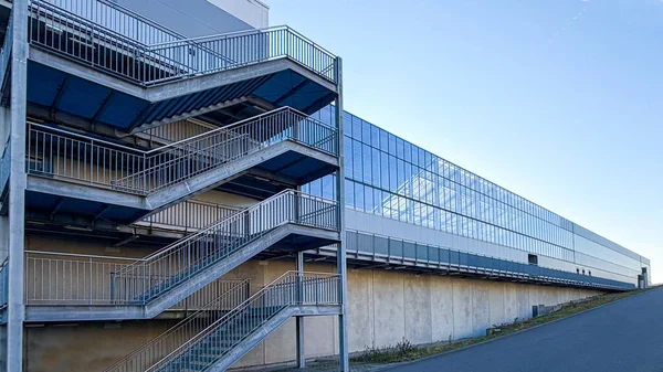 Dış cepheye bağlı metal bir merdiven bulunan sanayi deposu. Yapı beton duvarlar ve büyük cam yüzeyler, modern endüstriyel mimari, teknik altyapı ve lojistikle ilgili tesisleri birleştiriyor.