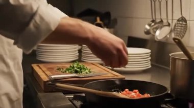Bir şef bir restoran mutfağında yemek hazırlar. Bir tava sebzeye doğranmış otlar ekliyor, sonra da onları tahta kaşıkla karıştırıyor. Tabaklar ve pişirme aletleri görünür..