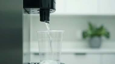 Suyun plastik bardağa döküldüğü yakın çekim videosu, bulanık bir mutfak arka planına ayarlanmış..