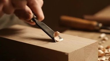 Bir ahşap işçisi tahta bloğu şekillendirmek için bir keski ve çekiç kullanır, odun kırıntıları yaratır. Eylem bir atölye ortamında gerçekleşiyor.