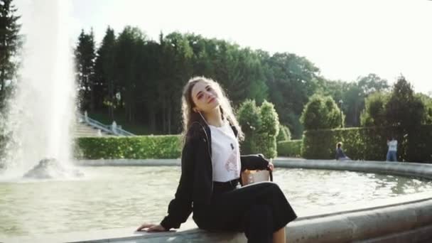 Jong Kaukasisch meisje ontspannen naast fontein in Europees park. Langzame beweging — Stockvideo