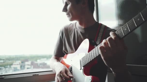 Молодий білий чоловік грає на червоній електрогітарі біля вікна в квартирі. Музичні вправи вдома. Повільні кадри руху — стокове відео