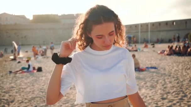 Curly Girl Girl regarde et sourit sur la plage de sable. Paysage européen ensoleillé. Joyeux jeune femme en t-shirt blanc. Millénaire en France. Look tendance dans un style rétro vintage. Caractère mignon — Video