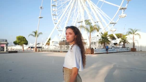 Curly Girl looks and enjoys the view. Sunny European landscape. Happy Young woman on the background of the Ferris wheel. Millennial in France. Cute character for travel blog or social media post. — Stockvideo