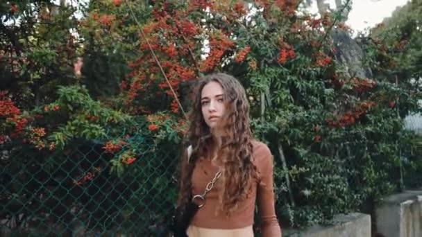 Ragazza riccia guarda e gode la vista. I capelli volano nel vento. Panorama europeo soleggiato. Giovane donna con un maglione beige per strada. Millennial sullo sfondo di alberi in Francia. Look alla moda. — Video Stock