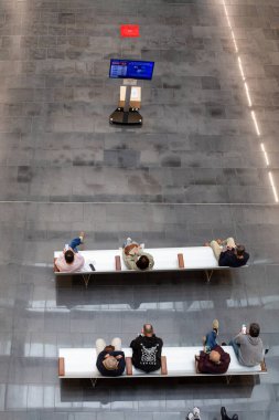 Hastalar ve ziyaretçiler temiz, minimalist bir hastane bekleme alanında sıralarda oturuyorlar, yukarıdan dijital bilgi ekranına bakıyorlar.