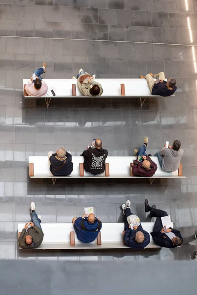 Modern bir hastane bekleme odasındaki banklarda oturan insanlar, cep telefonları kullanıyorlar ve üst perspektiften rahatlıyorlar.