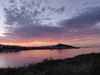 Gün batımı, ufukta bir kasaba ve tepe silueti olan sakin bir koyda canlı pembe ve mor yansımalar yaratır.