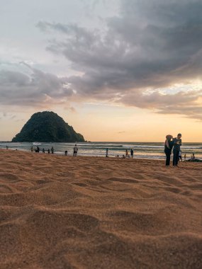 Pulau Merah (Red Island Beach), Banyuwangi 'deki kumlu plajın geniş açılı manzarasında ikonik yeşil kaplı adası yer almaktadır. İnsanlar, yumuşak ve bulutlu bir gökyüzünün altında rahatlar ve kıyı şeridi boyunca yürürler. Aile ve seyahat için mükemmel..