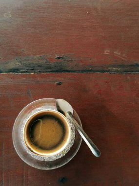 Koyu, ahşap bir masada gümüş kaşıkla servis edilen küçük, berrak bir cam fincanda servis edilen sıcak, koyu siyah bir kahvenin doğrudan yukarıdan görünüşü. Zengin kahverengi tonlar, desenli ahşap tahıl ve geniş kopya alanı var. Yemek, içecek ve yaşam tarzı için ideal..