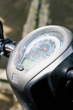 Tozlu, siyah scooter 'ın yuvarlak enstrüman kümesinin açılı yakın çekimi. Kırmızı aksanlı bir analog hız göstergesi ve aşağıda küçük, aydınlatılmamış bir dijital ekran özelliği.