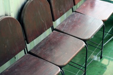 Çapraz çizgili basit, metal çerçeveli eski tahta sandalyeler, açık yeşil fayans zemin ve duvara karşı dizilmiş. Koyu renk ahşap ve aşınmış yüzeyler bir bekleme alanı, eski bir kafe ya da sınıf düzeni olduğunu gösteriyor..