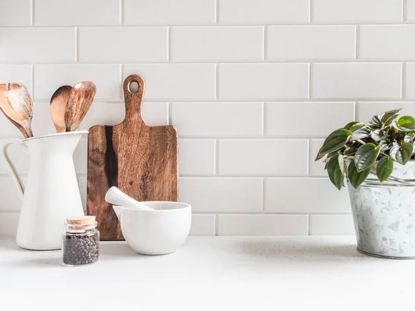 Stylish white kitchen background with kitchen utensils and green houseplant standing on white countertop, empty space for text, front view