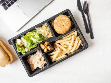 Pakette sezar salatası, hamburger ve patates kızartması olan öğle yemeği paketi paket servis edilecek veya yemek servisi ve dizüstü bilgisayar. Yemek teslimatı konsepti. Üst Manzara. Boşluğu kopyala