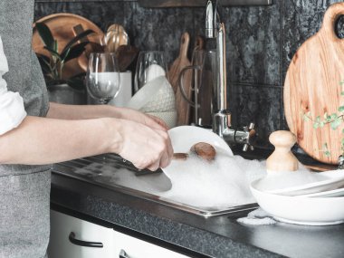 Gri önlüklü belirsiz bir kadın mutfakta lavabonun yanında ahşap fırçayla bulaşık yıkıyor. Ön görünüm