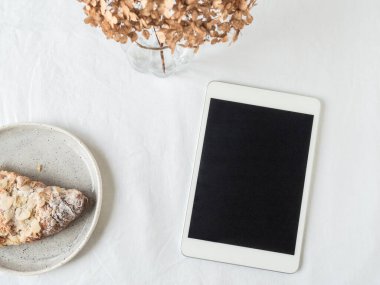 Boş siyah ekran tableti, kurumuş çiçekli cam vazo ve beyaz desenli arka planda kruvasan. Düz yatıyordu. Üst Manzara. Boşluğu kopyala