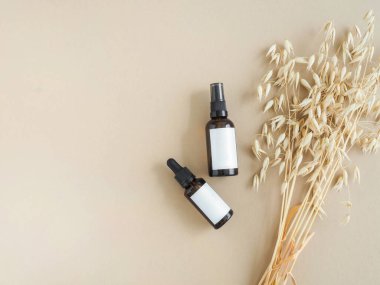 Bakım kozmetik ürünleri için iki kehribar şişesi, beyaz etiketli ve bej arka planda bir buket kuru yulaf. Üst Manzara. Boşluğu kopyala