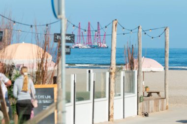 Kijkduin plajı, Lahey, Hollanda - 13 Haziran 2021: Hollanda açıklarında ağır endüstriyel rüzgar türbini teknesi olan sahil kafe, rüzgar enerjisi için bir açık deniz rüzgar parkı inşa ediyor.