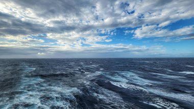 Dramatik bulutlu gökyüzü ve mavi bölgeler altında dinamik dalgalarla açık okyanus manzarası. Doğal deniz geçmişi, deniz yüzeyi dokusu, özgürlük, seyahat, macera ve hava durumu kavramı.