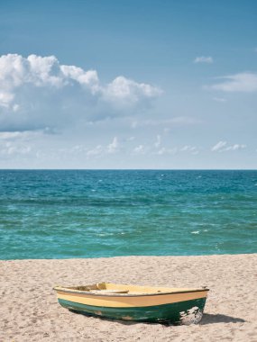 Açık mavi gökyüzünün altında turkuaz deniz kıyısında kumlu bir sahilde küçük sarı bir tekne.