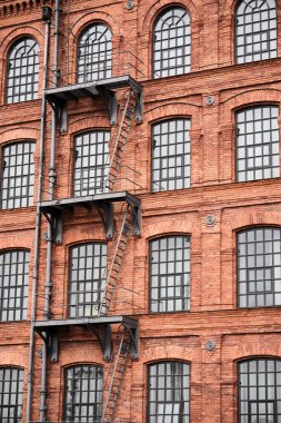 Kırmızı tuğlalı sanayi binasının ön cephesindeki çelik merdiven ve platformlar kemerli pencereler, mimari ve inşaat kavramları için güçlü dikey geometri.