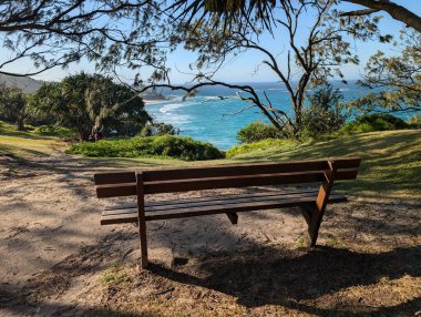 Kuzey Stradbroke Adası ahşap parkı. Çimenli sahil parkında parlak mavi okyanusa bakan bankta insanlar su kenarında dinleniyor. 