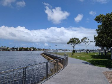 Kıvrımlı yürüme yolu, metal korkuluklar ve durgun su ve marinanın yanındaki yeşil park, açık mavi gökyüzünün altında güneşli bir günde tüylü beyaz bulutlarla birlikte..