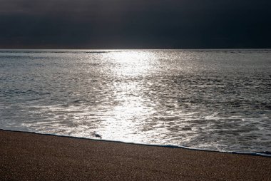 Okyanus sakin ve güneş suyun üzerinde parlıyor. Gökyüzü karanlık ve bulutlu. Gavres Yarımadası, Britanya, Fransa