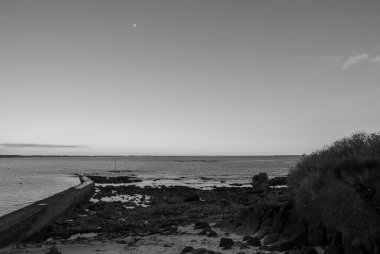 Görüntü, gökyüzünde büyük bir ay olan bir plajın siyah beyaz fotoğrafı. Sahil, ayın ışığını yansıtan suyla sakin ve huzurlu. Gavres Yarımadası, Britanya, Fransa
