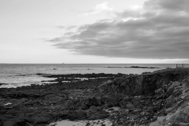 Arka planda bulutlu bir gökyüzü olan kayalık bir kıyı şeridi. Gökyüzü karanlık ve kayalar gri. Gavres Yarımadası, Britanya, Fransa