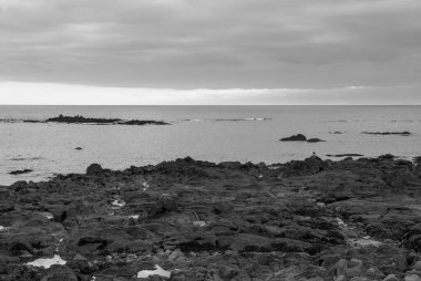 Arka planda sakin bir okyanus olan kayalık bir kıyı şeridi. Gökyüzü bulutlu, ortama dengesiz ve sakin bir atmosfer veriyor. Gavres Yarımadası, Britanya, Fransa