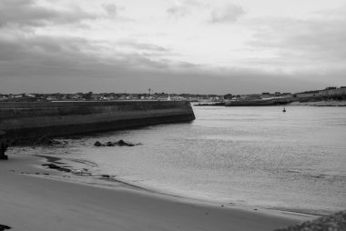 Görüntü, rıhtımı ve teknesi olan bir plajın siyah beyaz fotoğrafı. Sakin sular ve sakin bir ortam bir rahatlama ve huzur hissi yaratırken manzara sakin ve sakin. Gavres Yarımadası, Britanya, Fransa
